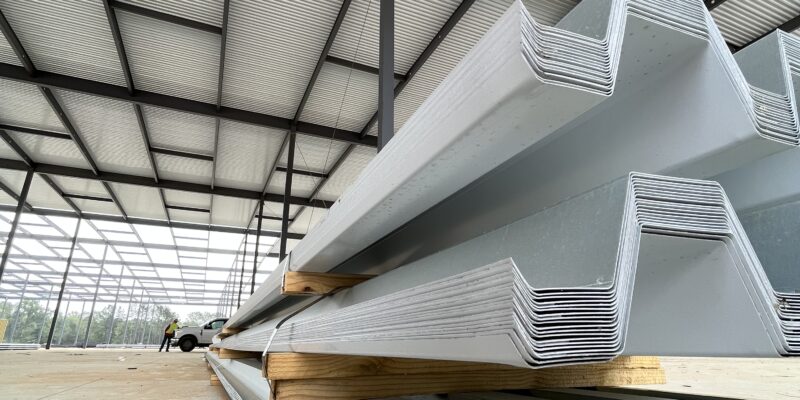 Stacked bundles of galvanized steel roof deck panels resting on wooden spacers inside a large steel-framed building, with exposed beams and a partially enclosed roof visible overhead.
